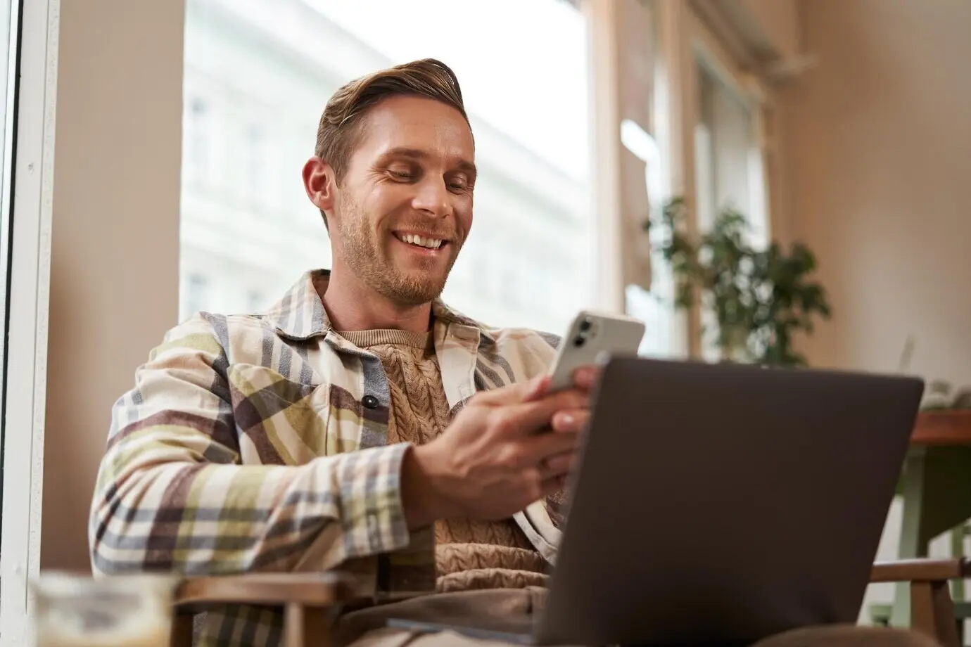 Крупный портрет привлекательного улыбающегося мужчины — посетителя кафе с ноутбуком, сидящего на стуле и пользующегося смартфоном.